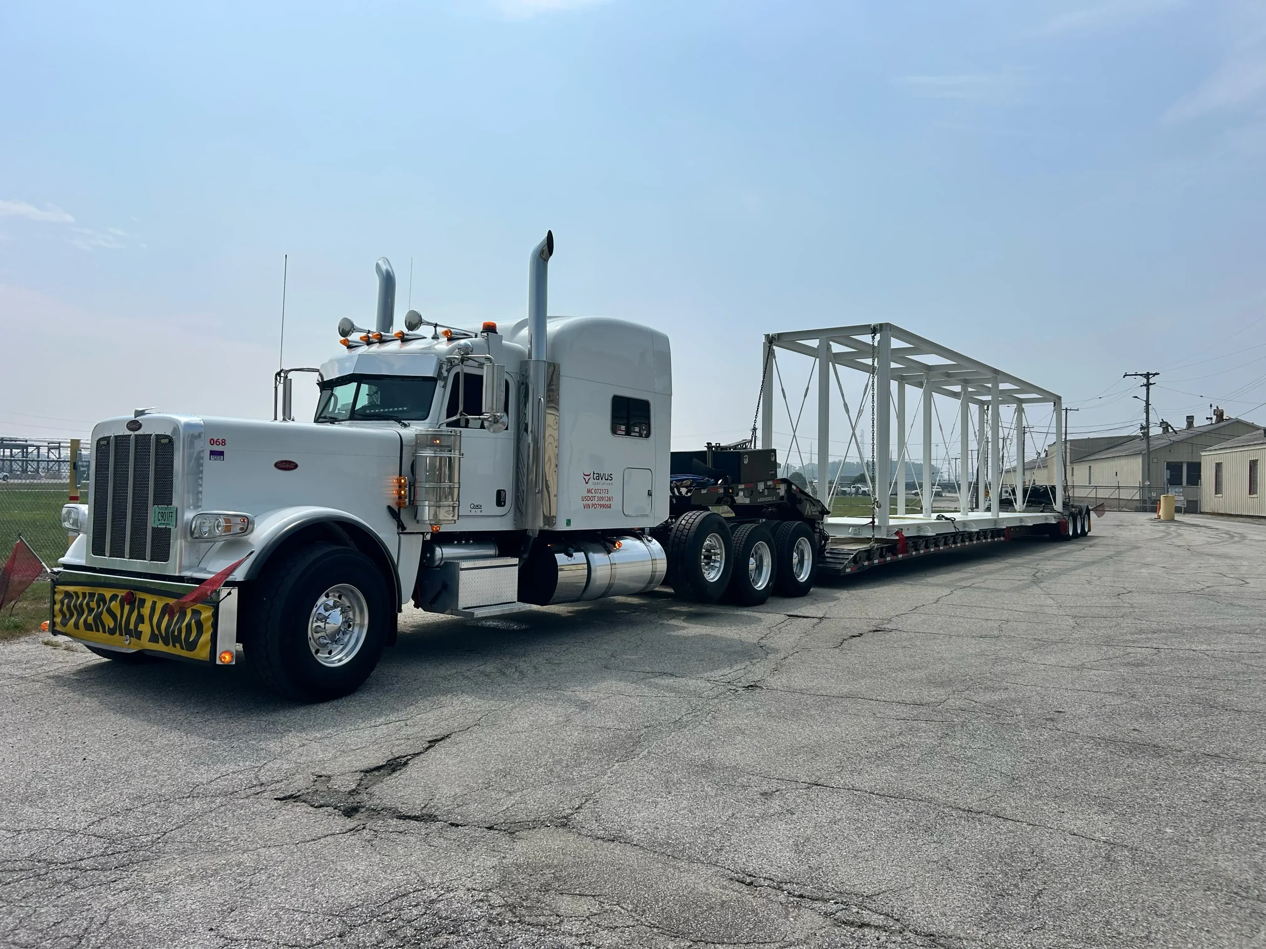 White Peterbilt with an oversize load banner hauling a steel truss frame on an RGN trailer